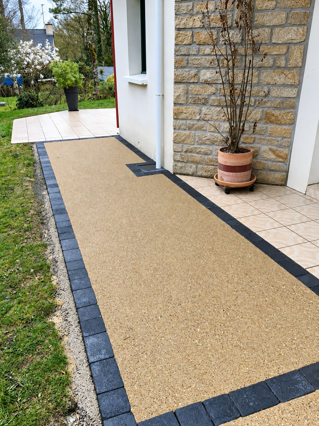 Allée étroite en béton micro-désactivé beige bordée de pavés anthracite, le long d'une façade en pierre et d'une pelouse.