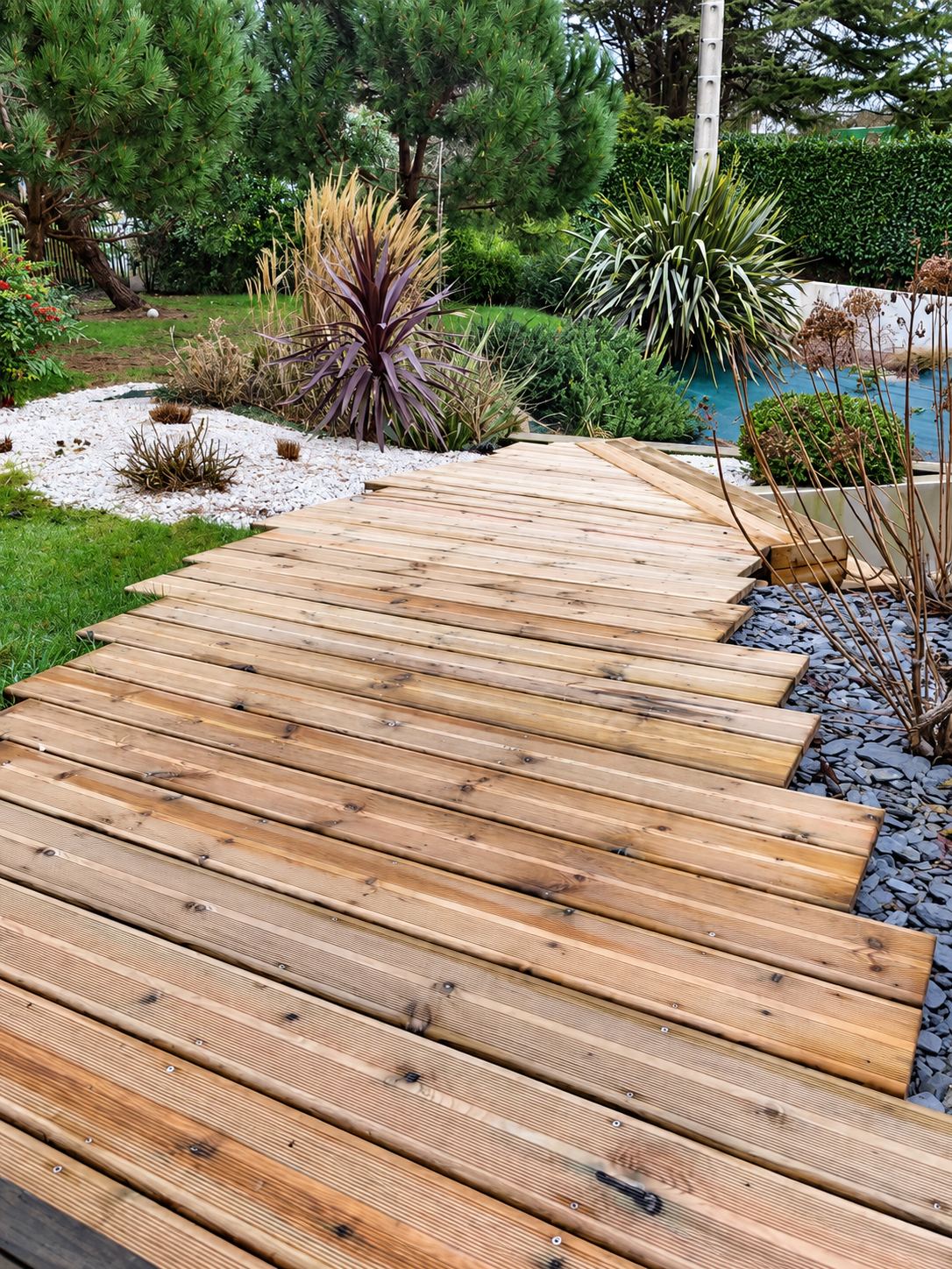 Une allée en lames de terrasse en bois serpente dans un jardin aménagé avec graviers blancs, ardoise bleue et plantes décoratives.
