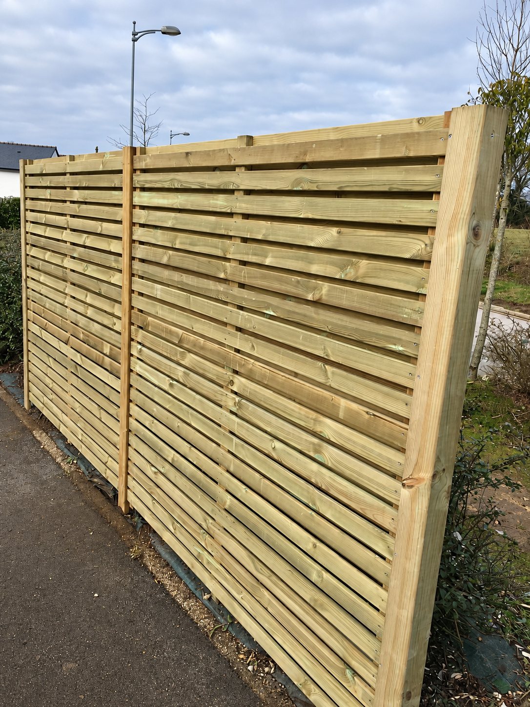 Une clôture en bois neuve à lames horizontales et poteaux carrés est installée le long d'un trottoir, avec un lampadaire et un ciel nuageux en arrière-plan.