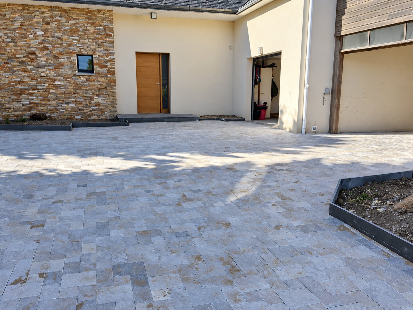 Une grande cour pavée de dalles de pierre grise devant une maison moderne avec un mur en pierre de parement et une porte en bois.