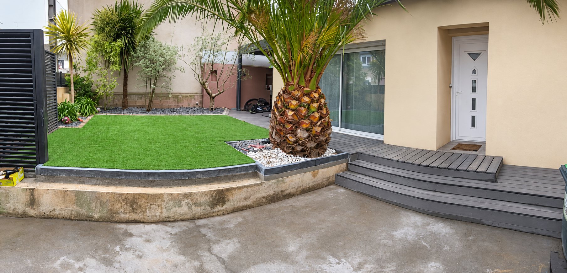 Petit jardin avec gazon synthétique, bordures en béton, palmier et terrasse devant une maison à façade claire.