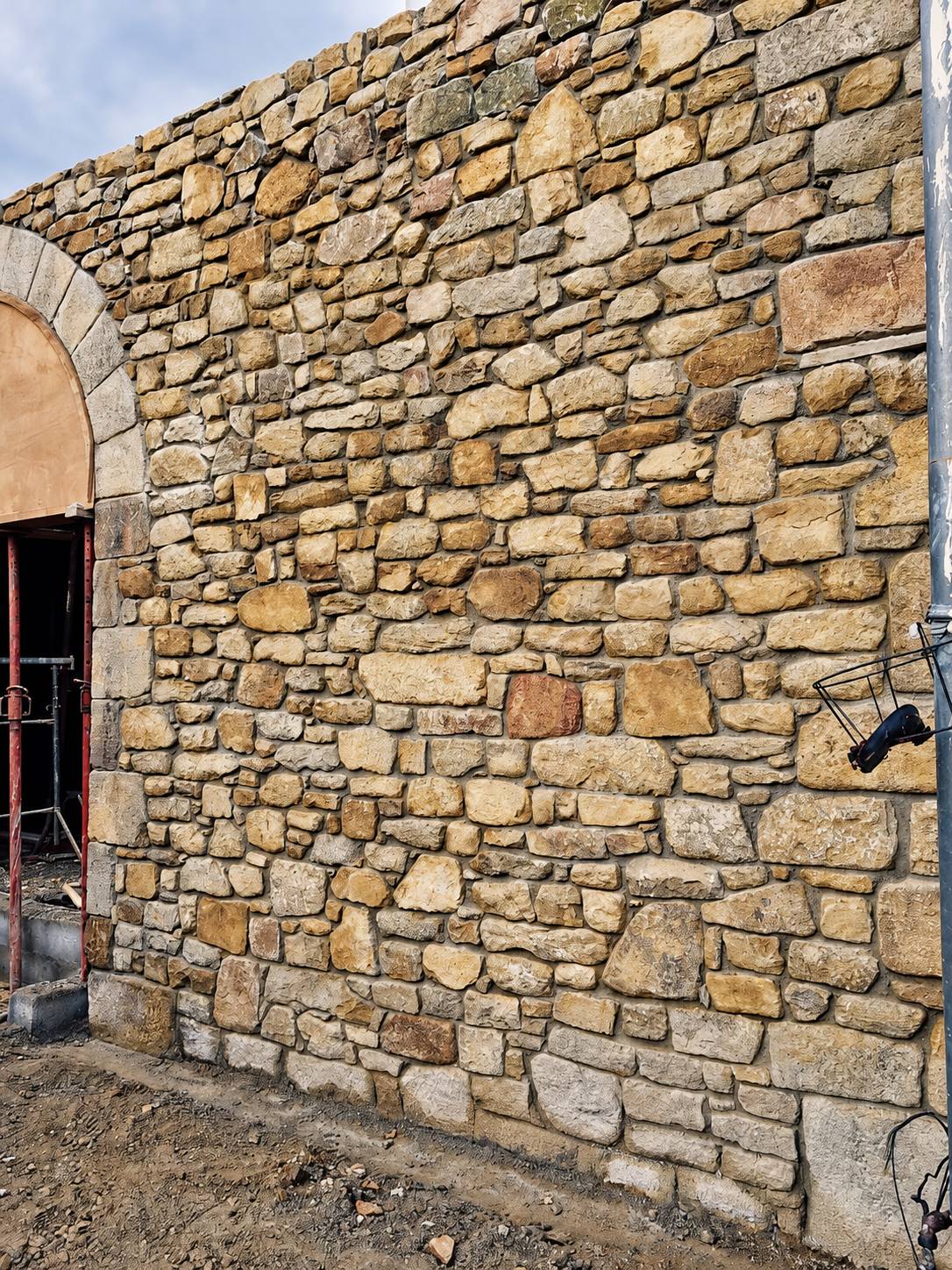 Mur en pierres apparentes dorées avec le départ d'une arche de porte et un échafaudage sur la gauche.