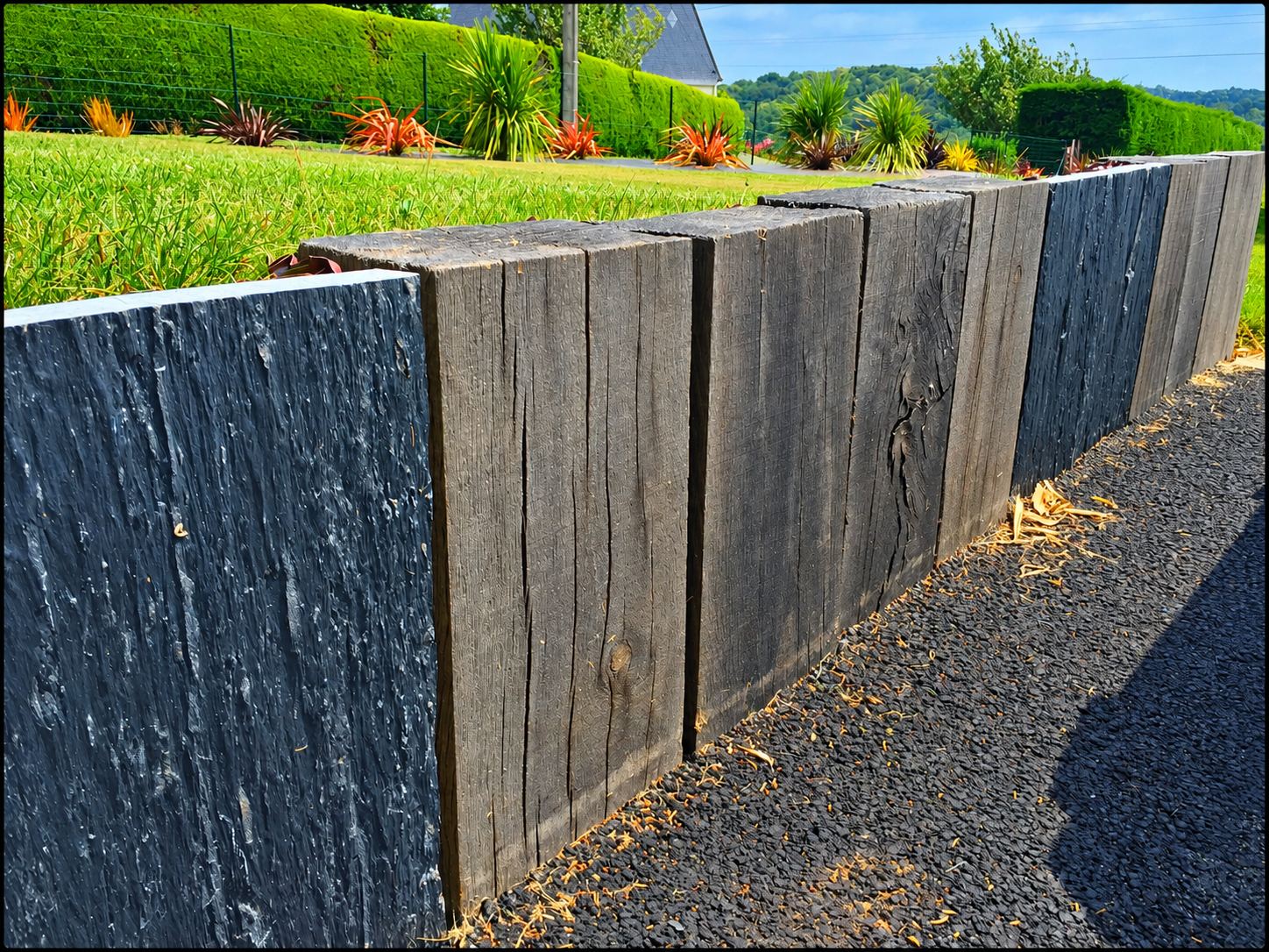 Un muret de jardin composé de traverses de bois sombre et de dalles d'ardoise borde une pelouse et des plantes ornementales, au-dessus d'une allée de gravillons noirs.