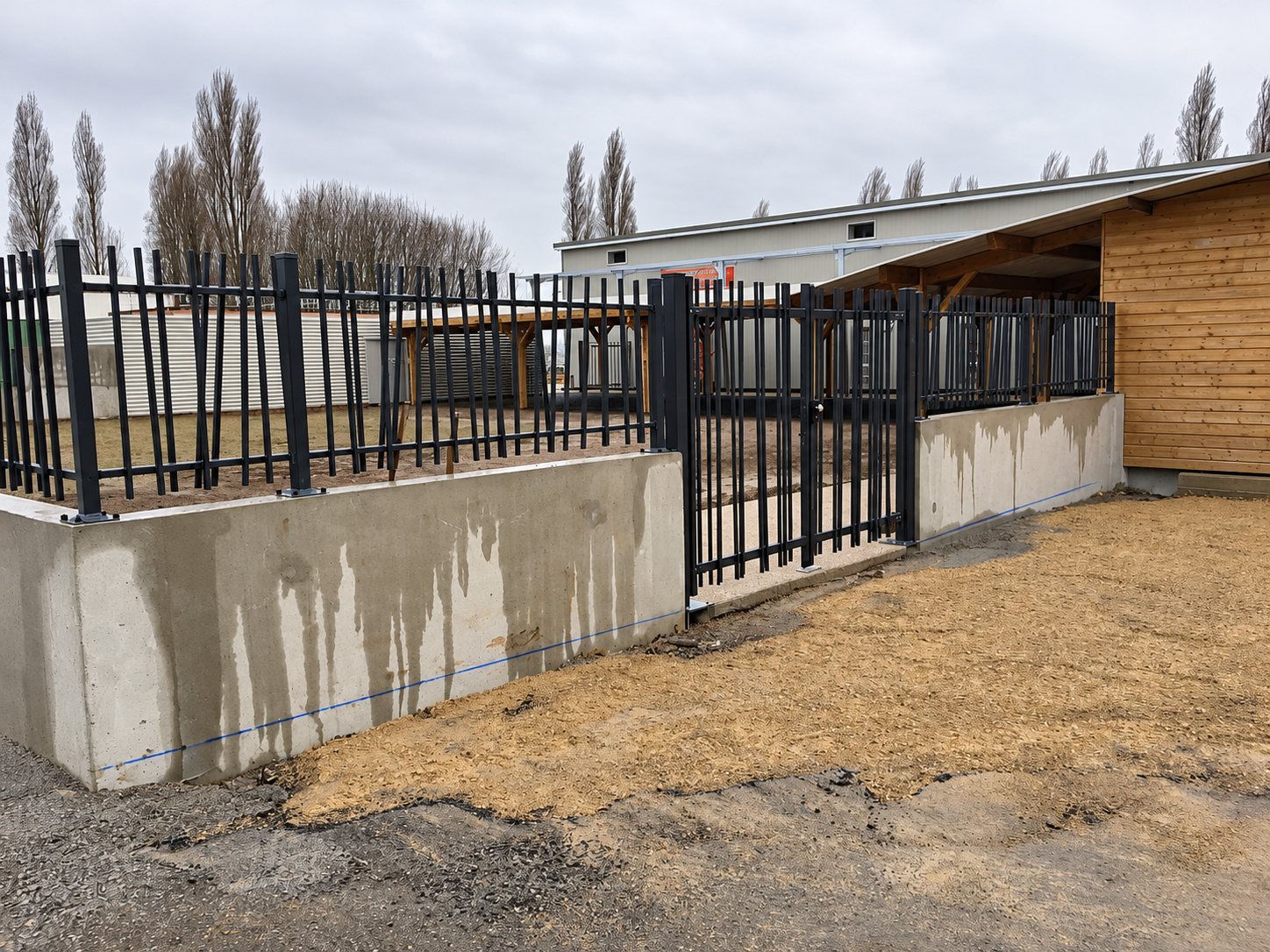 Clôture et portail à barreaudage noir posés sur un muret en béton, devant un bâtiment, avec de la terre nue au premier plan.