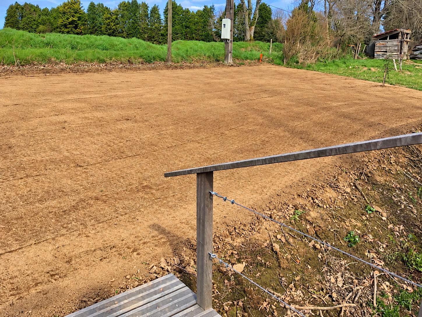 Une grande surface de terre nivelée et lissée en bordure d'un pré, avec une rambarde en bois et du fil de fer au premier plan.